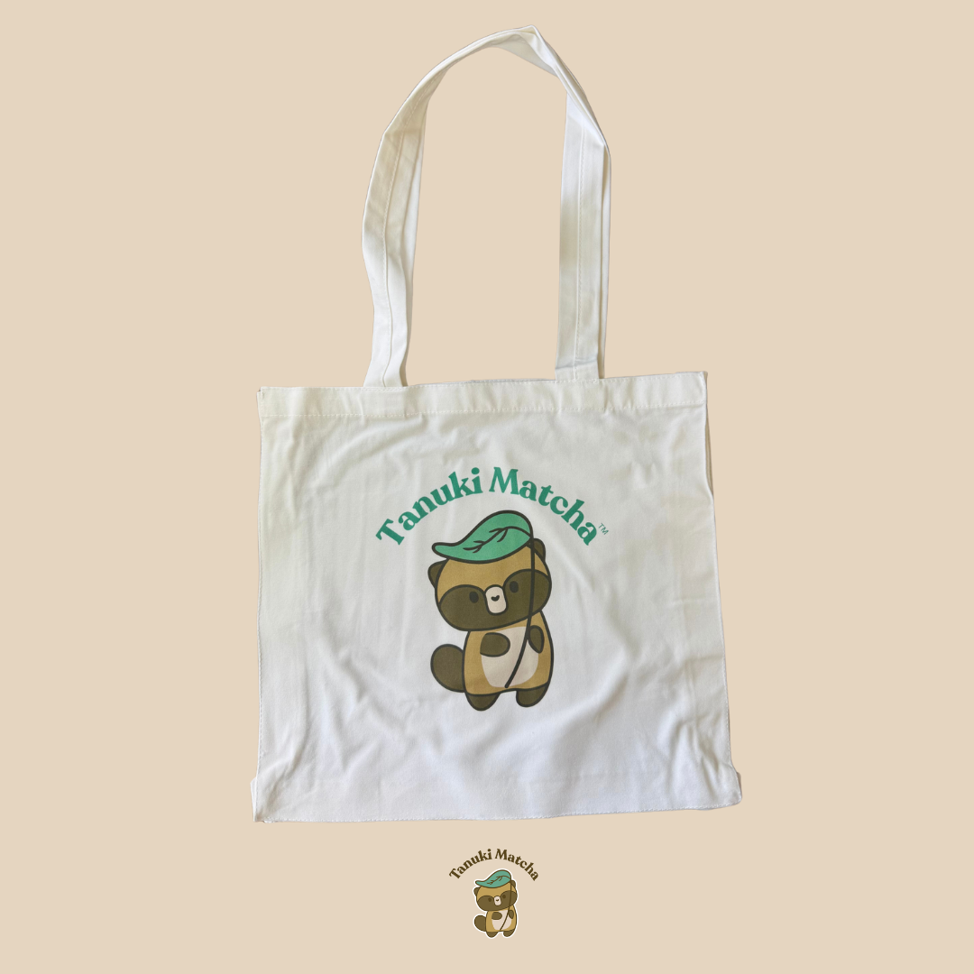 Tanuki Matcha Tote Bag. White tote bag with a cartoon raccoon and 'Tanuki Matcha' text on a beige background